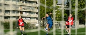 Durch das Netz eines Fußballtores sehen wir im Hintergrund eine Gruppe von Fußballspielerinnen.