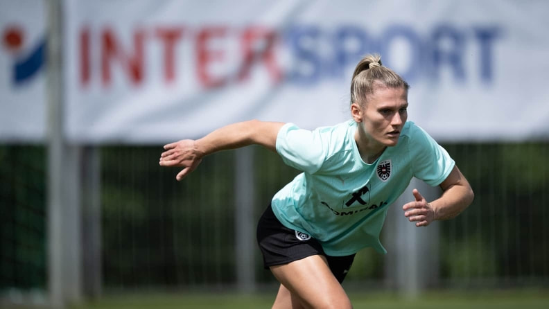 Sportlerin sprintet auf einem Fußballplatz mit nach vorne geneigtem Oberkörper und angewinkelten Armen. Im Hintergrund ist ein unscharfes Banner zu sehen.