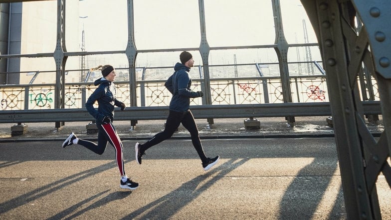 Zwei Leute joggen im Winter bei Sonnenschein über eine Brücke.