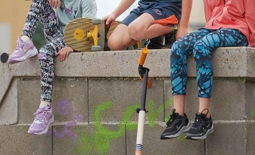 Drei Kinder sitzen auf einer Mauer mit einem Skateboard und einem Roller, ihre Beine in bunter Sportkleidung sichtbar.