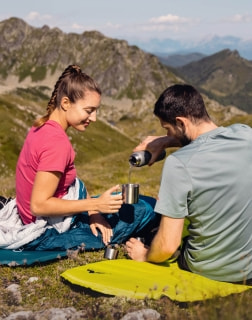 Frau und Mann sitzen auf Isomatten in den Bergen, während er aus einer Thermosflasche in einen Metallbecher einschenkt.