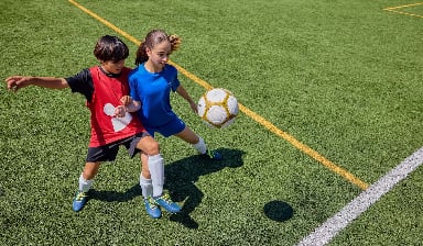 Ein Junge und ein Mädchen versuchen am Fußballfeld an den Ball zu kommen.