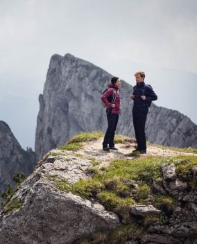 Zwei Wandernde stehen auf einem Felsvorsprung mit Rucksäcken vor steilen Berggipfeln.
