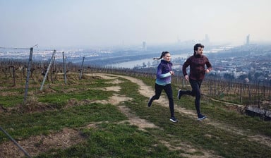 Ein Läufer und eine Läuferin laufen auf einem Hügel über einen Feldweg. Im Hintergrund sieht man im Tal eine große Stadt.
