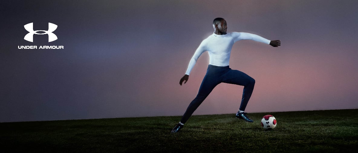 Under Armour Logo neben Fußballspieler in weißem Oberteil beim Fußballspielen auf einer Wiese