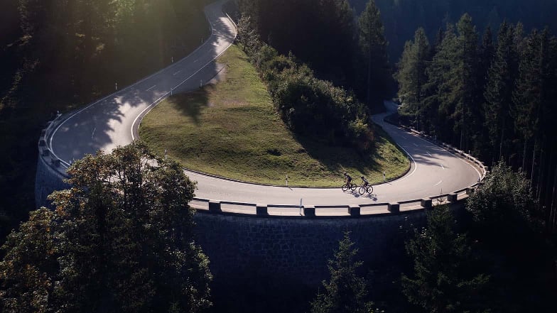 Die Kurve einer Bergstraße mit zwei Rennradfahrern in der Ferne.