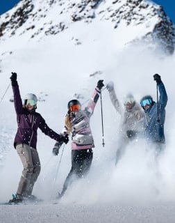 Vier Personen in Skiausrüstung jubeln gemeinsam im aufwirbelnden Schnee vor einer Bergkulisse.