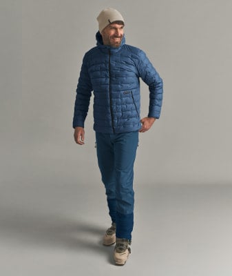 Ein Mann in einer blauen Daunenjacke und Skischuhen lächelt.