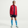 Mädchen in roter Jacke, dunkelblauen Shorts und hellblauen Socken.