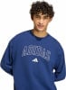 Ein Mann mit lockigem Haar trägt einen blauen Pullover mit Adidas-Logo.