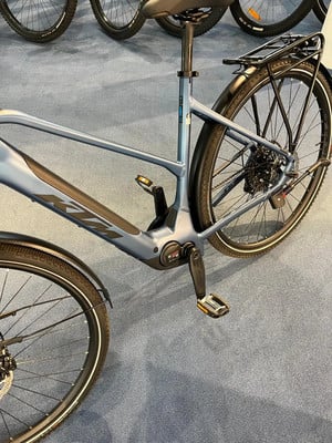 Ein blaues E-Bike mit Gepäckträger und Schutzblechen auf grauem Teppich.