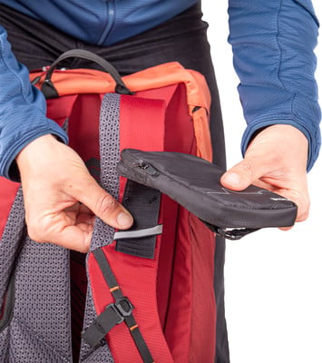 Eine Hand hält eine kleine schwarze Tasche mit Reißverschluss an einem roten Rucksack.