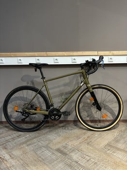 Ein grünes Gravelbike steht auf einem Holzboden vor einer grauen Wand.