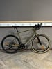 Ein grünes Gravelbike steht auf einem Holzboden vor einer grauen Wand.