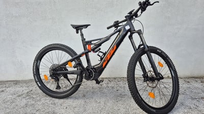 Ein schwarzes KTM E-Mountainbike mit orangefarbenen Akzenten steht auf Schotter. Ein schwarzes KTM E-Mountainbike mit orangefarbenen Akzenten steht auf Schotter.