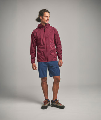Ein Mann in einer dunkelroten Jacke und blauen Shorts steht vor grauem Hintergrund.