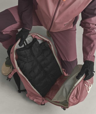 Ein rosafarbener Rucksack mit schwarzer Daunenjacke, Hände in Handschuhen packen.