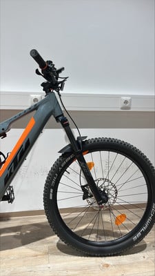 Vorderseite eines grauen E-Mountainbikes mit orangefarbenen Akzenten und Federung. Vorderseite eines grauen E-Mountainbikes mit orangefarbenen Akzenten und Federung.