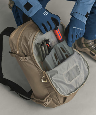 Eine Person verstaut einen orangefarbenen Stab und Skistöcke in einem Rucksack.