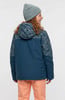 Mädchen in blauer Jacke mit Muster und orangefarbener Hose trägt eine Mütze.