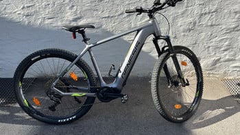 Ein graues Genesis E-Bike mit schwarzen Rädern und gelben Akzenten steht an einer Wand.