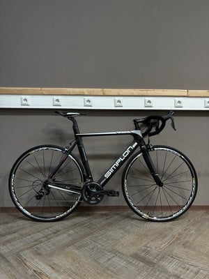 Ein schwarzes Simplon Rennrad steht auf einem Holzboden vor einer grauen Wand. Ein schwarzes Simplon Rennrad steht auf einem Holzboden vor einer grauen Wand.