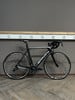 Ein schwarzes Simplon Rennrad steht auf einem Holzboden vor einer grauen Wand.