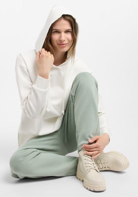 Eine Frau in einem weißen Hoodie und grüner Jogginghose sitzt auf dem Boden.