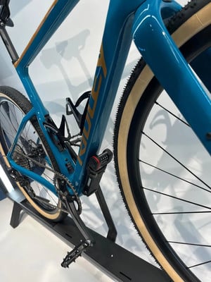 Blauer Gravelbike mit braunen Details und schwarzen Pedalen auf einem Ständer.
