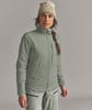 Eine Frau in einer beige Mütze und grüner Jacke steht vor grauem Hintergrund.