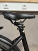 Schwarze Sattelstütze mit schwarzem Sattel auf einem Fahrrad, Steckdosen im Hintergrund.
