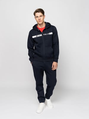 Egy fiatalember sötétkék melegítőben és fehér sportcipőben pózol fehér háttér előtt.