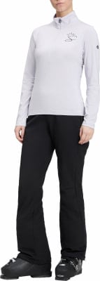 Eine Frau in Skikleidung, weißes Oberteil und schwarze Hose, steht in Skischuhen.