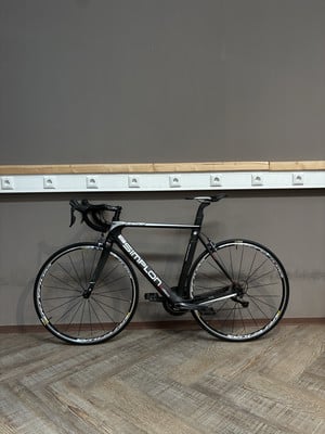 Ein schwarzes Simplon Rennrad steht auf einem Holzboden vor einer grauen Wand. Ein schwarzes Simplon Rennrad steht auf einem Holzboden vor einer grauen Wand.