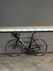 Ein schwarzes Simplon Rennrad steht auf einem Holzboden vor einer grauen Wand.