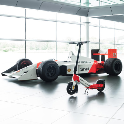 Ein McLaren-Rennwagen und ein Elektroroller stehen nebeneinander in einer Halle. Ein McLaren-Rennwagen und ein Elektroroller stehen nebeneinander in einer Halle.