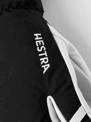 Detail eines Handschuhs mit HESTRA-Logo, schwarzer Stoff und weißes Leder. Detail eines Handschuhs mit HESTRA-Logo, schwarzer Stoff und weißes Leder.