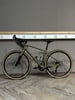 Ein grünes Gravelbike steht auf einem Holzboden vor einer grauen Wand.