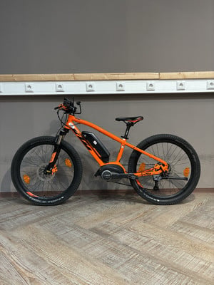 Ein orangefarbenes E-Bike mit schwarzen Rädern steht auf einem Holzboden. Ein orangefarbenes E-Bike mit schwarzen Rädern steht auf einem Holzboden.