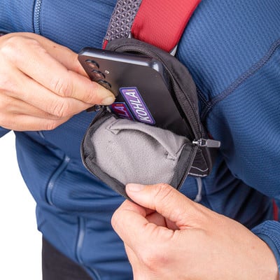 Eine Hand steckt ein Handy in eine kleine schwarze Tasche auf der Brust eines Rucksacks.