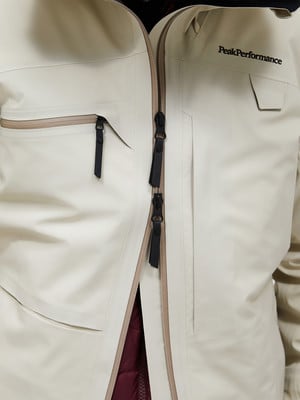 Eine beige Jacke mit schwarzem Reißverschluss und Peak Performance Logo. Eine beige Jacke mit schwarzem Reißverschluss und Peak Performance Logo.