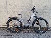 Silbernes E-Bike mit Gepäckträger und Schutzblechen vor Betonwand.