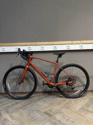 Ein orangefarbenes Gravelbike steht auf einem Holzboden vor einer grauen Wand. Ein orangefarbenes Gravelbike steht auf einem Holzboden vor einer grauen Wand.