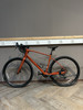 Ein orangefarbenes Gravelbike steht auf einem Holzboden vor einer grauen Wand.