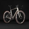 Ein beiges Gravelbike mit schwarzem Lenker und braunen Reifen vor schwarzem Hintergrund.