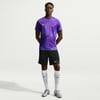 Ein Mann in einem lila Hemd mit geometrischem Muster und schwarzen Shorts.