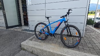 Ein blaues Mountain-E-Bike steht vor einem Gebäude mit grauen Wänden.