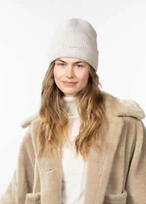Eine Frau in einem beigen Teddyfellmantel und einer hellbeigen Mütze mit Logo. Eine Frau in einem beigen Teddyfellmantel und einer hellbeigen Mütze mit Logo.