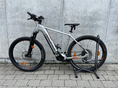 Ein silbernes E-Mountainbike mit schwarzen Rädern vor einer Betonwand.