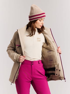 Eine Frau in einer beigen Jacke und rosa Hose, mit Mütze auf dem Kopf.